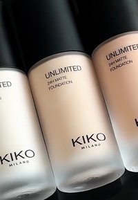 Tre frostat glasflaskor med KIKO Milano Unlimited 24H Matte Foundation, med svarta lock och genomskinliga etiketter som anger produktens namn.