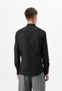 Homme portant une chemise noire à manches longues et un pantalon gris, debout de dos sur un fond blanc uni.