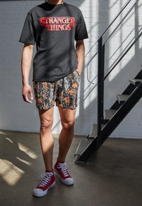 Persona con camiseta negra de "Stranger Things", pantalones cortos florales y zapatillas rojas de caña alta, de pie cerca de una escalera de metal en una habitación iluminada por el sol.