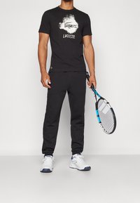 Mies, jolla on päällään musta Lacoste-t-paita ja mustat housut, pitää mustaa ja sinistä tennismailaa käsissään, seisten yksinkertaisella taustalla.