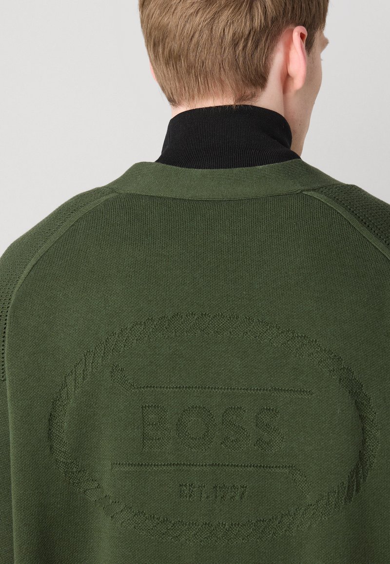 Zielony sweter robiony na drutach z czarnym ściągaczem. Posiada wytłoczone logo na plecach. Gładka tekstura z subtelnymi detalami wzdłuż szwów.