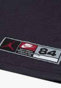 Μαύρη υφασμάτινη ετικέτα με γκρίζες, κόκκινες και λευκές λεπτομέρειες. Περιλαμβάνει το λογότυπο "Jordan", το κείμενο "Nike" και τον αριθμό "84", με περιγραφικό κείμενο από κάτω.