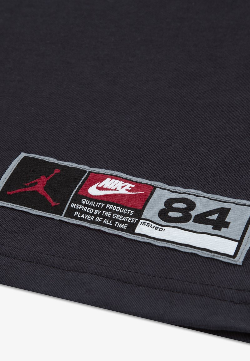 Μαύρη υφασμάτινη ετικέτα με γκρίζες, κόκκινες και λευκές λεπτομέρειες. Περιλαμβάνει το λογότυπο "Jordan", το κείμενο "Nike" και τον αριθμό "84", με περιγραφικό κείμενο από κάτω.