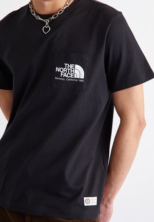 Person iført sort The North Face-T-shirt med hvidt logo og tekst på brystlommen samt en halskæde med et hjerteformet vedhæng.