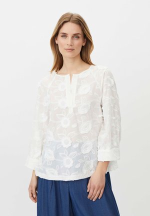 Kvinde med lige lys brun hår iført en hvid blomstret broderet bluse med lange ærmer og løse blå bukser, stående mod en ensfarvet baggrund.
