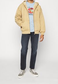Sudadera con cremallera beige con un forro de felpa texturizado, llevada sobre una camiseta azul claro con un estampado gráfico, combinada con unos vaqueros oscuros y zapatillas de cuadros.
