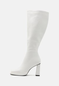 Botte blanche jusqu'au genou en matériau lisse, dotée d'un talon haut épais et d'un embout légèrement carré avec des accents noirs sur la semelle.