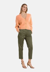 Perzikkleurige gebreide cardigan met een V-hals en gouden knopen, gecombineerd met olijfgroene broek en bronzen sandalen met bandjes.