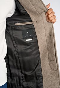 Hånd, der holder en åben beige uldfrakke, der viser sort foring med "JUNK de LUXE" mærke og indre lomme.