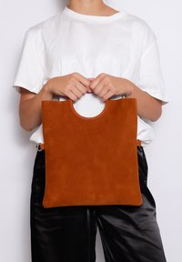 Borsa tote in suede color ruggine con manico a forma di cerchio. Presenta un design semplice e rettangolare, con una texture liscia e senza accenti hardware visibili.
