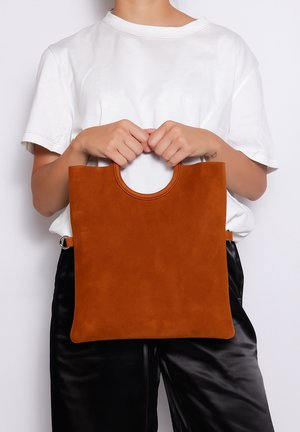 Suède tote bag in roestkleur met een ronde uitsnijding als handvat. Heeft een eenvoudig, rechthoekig ontwerp en een gladde textuur, zonder zichtbare hardware accenten.