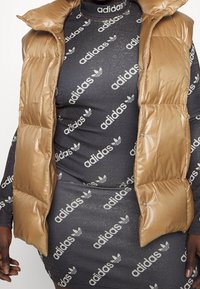 Doudoune sans manches matelassée de couleur marron clair avec une texture brillante, portée sur une robe ajustée gris foncé ornée de logos Adidas blancs répétés.