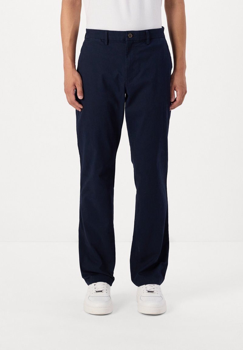 Homme portant un pantalon droit bleu marine et des baskets blanches, debout devant un fond clair uni.