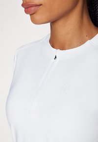 Top atlético blanco con cuello con cremallera, que presenta un logo sutil y una tela suave y ligera. La vista en primer plano destaca los detalles de la costura.