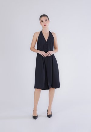 Nainen seisoo yksivärisen taustan edessä pukeutuneena hihattomaan mustaan polvipituiseen mekkoon ja mustiin korkeakorkoisiin kenkiin, kädet ristissä edessä.