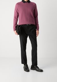 Personne portant un pull violet à côtes avec un col noir, un pantalon noir et des chaussures noires à lacets, tenant une pochette en cuir noir.