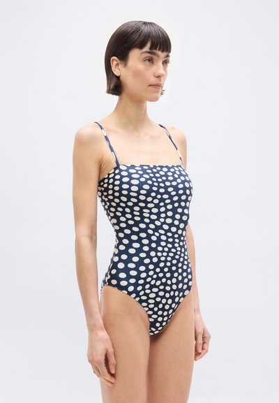 Lühikeste tumejuustega naine, kes kannab mereväesinist üheosalist ujumistrikood beežide ebaühtlaste täppidega, seisab valge tausta ees.