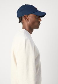 Porträt eines Mannes, der eine marineblaue Baseballmütze und ein off-white Langarmhemd vor einem schlichten hellen Hintergrund trägt.