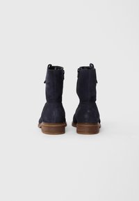 Bottines en daim bleu marine avec une texture lisse, bout rond et talon en bois. Elles disposent d'une fermeture éclair sur le côté et d'une boucle à l'arrière pour un enfilage facile.