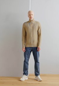 Maglione beige con zip al collo e texture a onda, abbinato a jeans blu e sneakers bianche con dettagli crema. Design semplice e moderno.