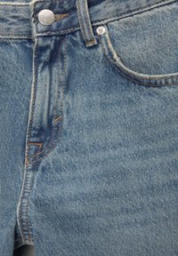 Gros plan sur un jean en denim bleu montrant la poche avant, le bouton en métal et les coutures détaillées sur le tissu texturé.