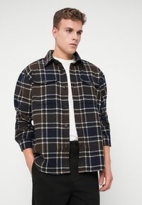 Camicia in flanella a quadri marrone e blu navy con chiusura a bottoni, due tasche sul petto e colletto a punta. Maniche lunghe, vestibilità rilassata.