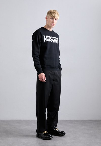 Ung man med blont hår, iklädd en svart Moschino-tröja, lösa svarta byxor och svarta skor med guld detaljer, stående på ett grått golv.
