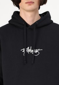 Crna dukserica s kapuljačom na vezanje i bijelim izvezbenim logom na prsima. Izrađena je od mekane, teksturirane tkanine.