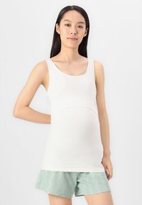 Λευκή αμάνικη μινι φόρμα εγκυμοσύνης με στρογγυλή λαιμόκοψη και μαζεμένο ύφασμα στην κοιλιά. Συνδυασμένη με ελαφριά πράσινα υφασμάτινα σορτς.