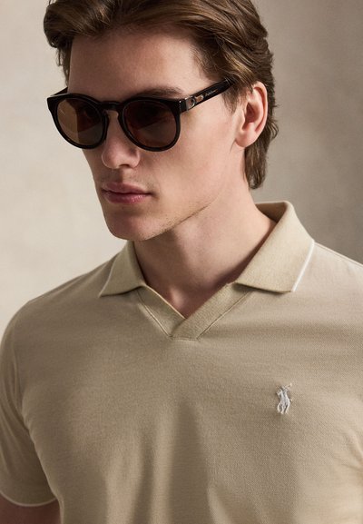Νεαρός άνδρας με σκούρα καφέ γυαλιά ηλίου και μπεζ πόλο με μικρό λευκό κεντητό λογότυπο στο στήθος.