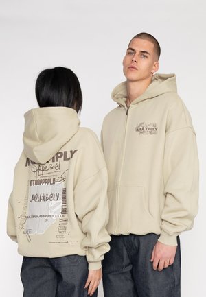 Sudadera con cremallera en color beige, con un gran bolsillo delantero y un ajuste oversize. Presenta gráficos impresos en la parte posterior y en el pecho en tonos marrones apagados.