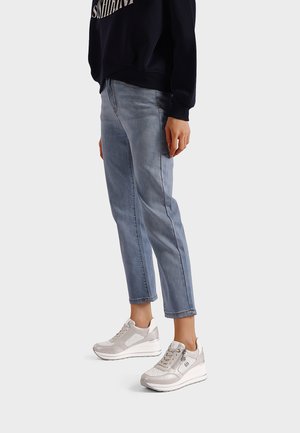 Persona che indossa jeans corti azzurri chiari e sneakers bianche con piattaforma e zip laterali, in piedi contro uno sfondo semplice.