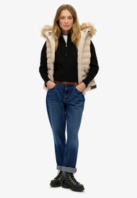 Beige gewatteerde vest met een bont-gevoerde capuchon, gedragen over een zwarte kabelgebreide trui. Blauwe opgerolde jeans en zwarte veterboots maken de look compleet.