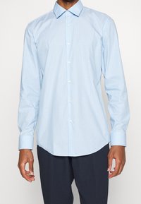 Uomo che indossa una camicia da uomo azzurra chiara a righe sottili con maniche allacciate e pantaloni scuri, in piedi davanti a uno sfondo neutro.