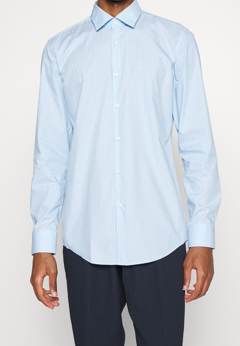 Uomo che indossa una camicia da uomo azzurra chiara a righe sottili con maniche allacciate e pantaloni scuri, in piedi davanti a uno sfondo neutro.