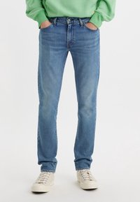 Personne portant un jean bleu moyen à jambes droites, des baskets blanches et un sweat-shirt vert avec les mains dans les poches, debout sur un fond uni.