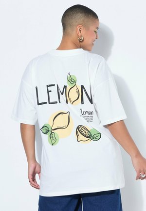 Person mit kurzen Haaren, trägt ein weißes, übergroßes T-Shirt mit Zitronenillustrationen und dem Text "LEMON" auf der Rückseite, kombiniert mit blauen Hosen.