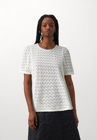 Hvit blondebluse med et teksturert diamantmønster, korte puffermer og rund hals, kombinert med et mørkt quiltet skjørt.
