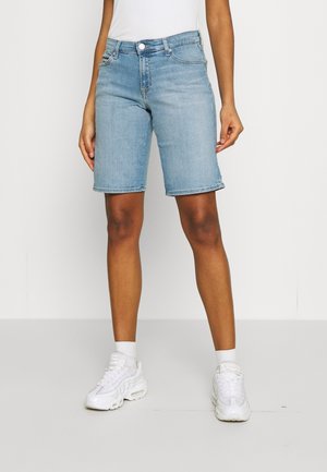 Personne portant un short en jean bleu clair longueur genou, des chaussettes blanches et des baskets blanches, debout devant un fond uni.