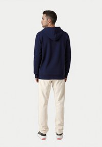 Felpa blu navy con cappuccio doppio, polsini e orlo a costine, abbinata a pantaloni chiari e scarpe scure con suola bianca.