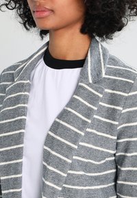 Grå och vit randig blazer med flikkrage, texturerad tyg och öppen design, lager på lager över en svart och vit topp.