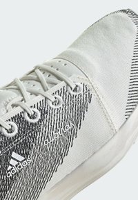 Zapatilla deportiva blanca con una parte superior texturizada, rayas diagonales negras, costuras reforzadas y un branding destacado en el lado.