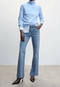 Un pull col roulé bleu clair, une chemise blanche à col en dessous, et un jean bleu clair évasé avec des bords bruts, associés à des escarpins noirs à bout pointu.