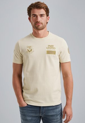 Camiseta de manga corta de color beige claro con textura acanalada, que presenta logos y parches bordados en dorado en el pecho y la manga.
