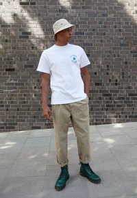 Camiseta blanca de algodón con gráfico azul, pantalones tapered beige y botas de cordones verde oscuro con suelas texturizadas, frente a un fondo de ladrillo.