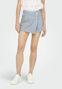 Mini-jupe en denim bleu clair avec une taille haute, fermeture à boutons sur le devant et poches discrètes. Associée à des baskets blanches pour un look décontracté.