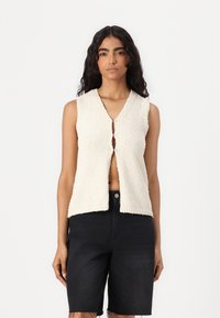 Gilet sans manches texturé beige avec un col en V et une fermeture à boutons sur le devant, associé à un short en denim noir. Pas de motifs ni d'accents visibles.