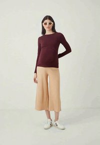 Μακρυμάνικο τοπ εγκυμοσύνης σε μπορντώ απόχρωση, συνδυασμένο με μπεζ πλουσιές παντελόνες. Το μοντέλο φοράει λευκά αθλητικά παπούτσια και oversized γυαλιά ηλίου.