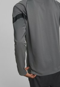 Tummanharmaa pitkähihainen urheilupaita, jossa on teksturoitu kangas ja musta korostevyö olkapäällä, jossa pienet rei'itykset.