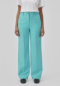 Pantalons larges turquoise en tissu lisse, avec des plis sur le devant et une fermeture à bouton, associés à un t-shirt blanc uni.